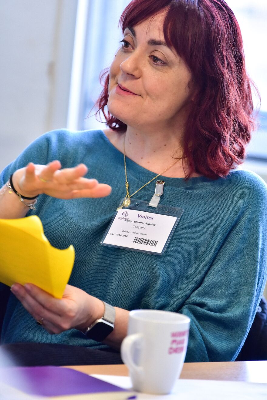 A woman with red hair and wearing a blue jumper holding a bright yellow piece of paper.